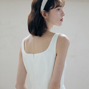 May include: A white sleeveless dress with a square neckline and a back zipper. The dress is fitted through the bodice and flows out at the hem. The model is wearing a white headband with pearl embellishments.