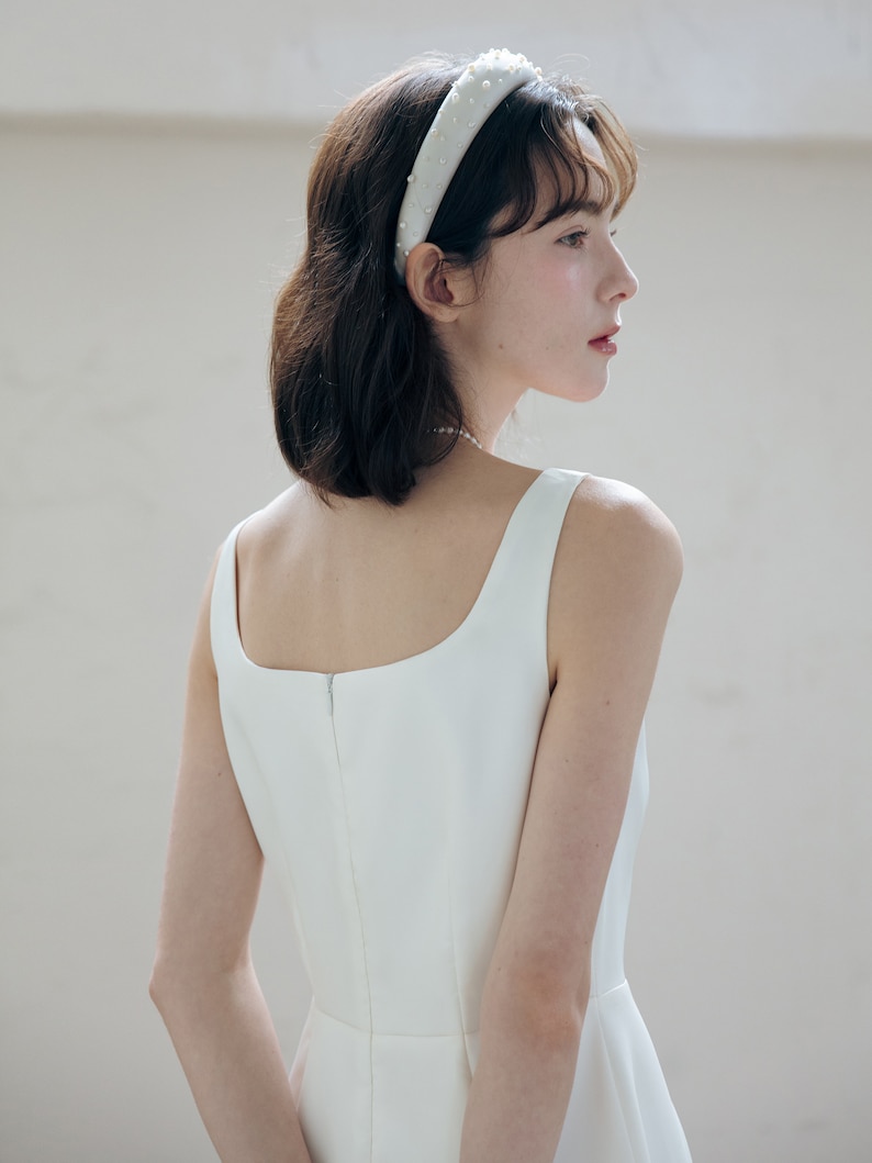 May include: A white sleeveless dress with a square neckline and a back zipper. The dress is fitted through the bodice and flows out at the hem. The model is wearing a white headband with pearl embellishments.