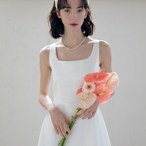 May include: A white sleeveless dress with a square neckline and a flowing skirt. The dress is paired with a white pearl headband and a bouquet of pink and orange flowers.