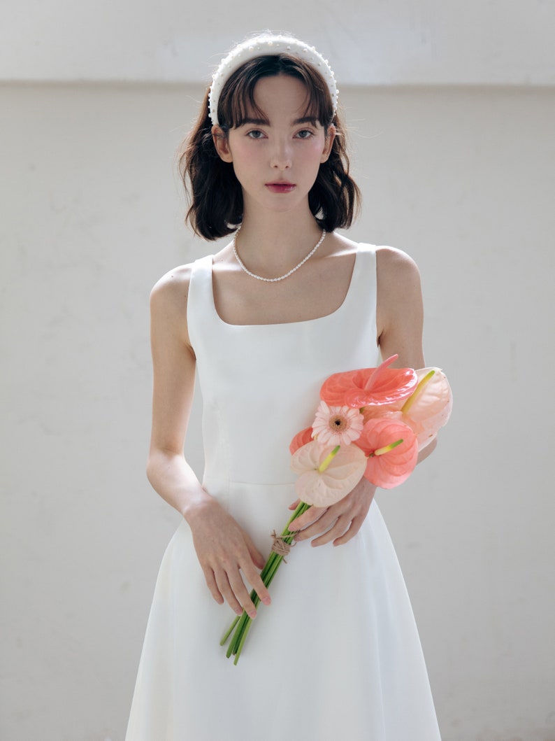 May include: A white sleeveless dress with a square neckline and a flowing skirt. The dress is paired with a white pearl headband and a bouquet of pink and orange flowers.