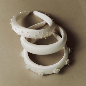 May include: Three white satin headbands with pearl and floral embellishments. The headbands are stacked on top of each other.