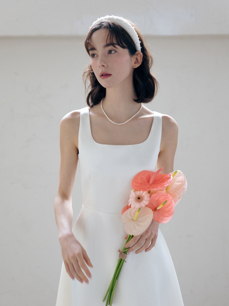 May include: A woman wearing a white sleeveless dress with a pearl necklace and a white headband with pearl accents. She is holding a bouquet of pink and white flowers.