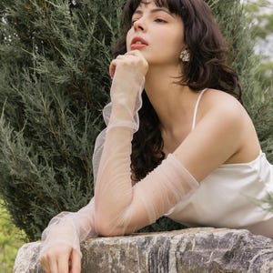 May include: A woman wearing a white sleeveless top and sheer white gloves, resting her chin on her hand, looking up and to the right. She is sitting on a large, gray rock with green foliage behind her.