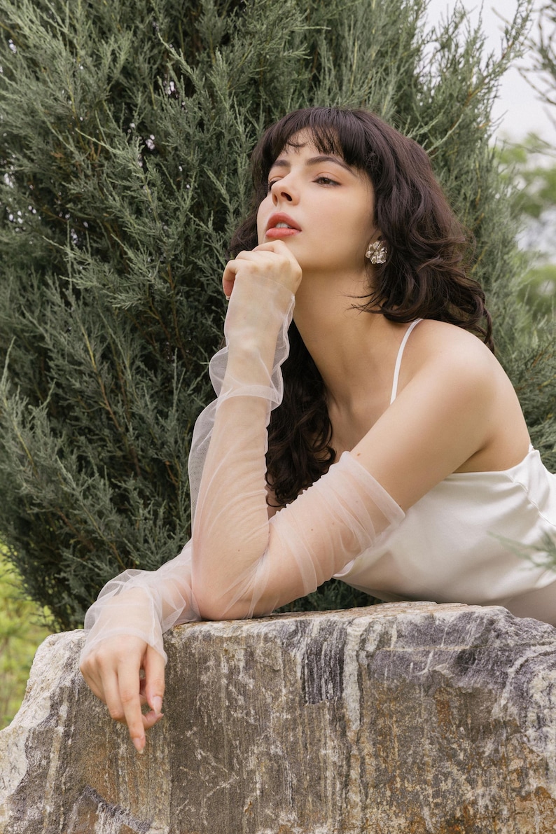 May include: A woman wearing a white sleeveless top and sheer white gloves, resting her chin on her hand, looking up and to the right. She is sitting on a large, gray rock with green foliage behind her.