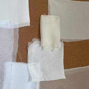 May include: Close-up of a corkboard with several pieces of white fabric pinned to it. The fabrics vary in texture and opacity, ranging from sheer to opaque. The pins are silver and hold the fabric in place.