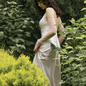 May include: A woman in a white satin dress with sheer sleeves stands in a garden setting. The dress has a simple, elegant design and flows gracefully around her.