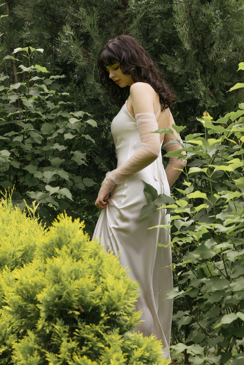 May include: A woman in a white satin dress with sheer sleeves stands in a garden setting. The dress has a simple, elegant design and flows gracefully around her.
