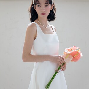 May include: A white sleeveless wedding dress with a square neckline and a flowing skirt. The bride is wearing a white pearl headband and a pearl necklace. She is holding a bouquet of pink flowers.