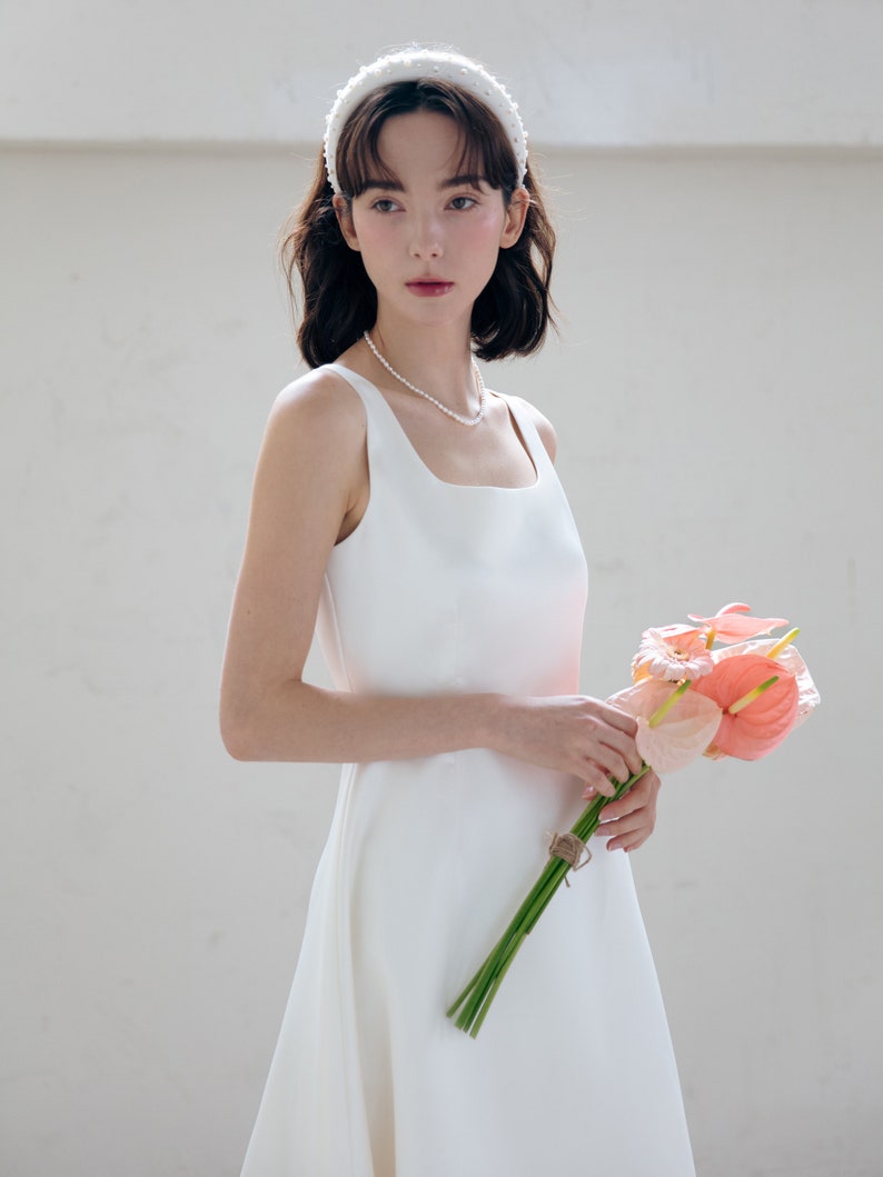 May include: A white sleeveless wedding dress with a square neckline and a flowing skirt. The bride is wearing a white pearl headband and a pearl necklace. She is holding a bouquet of pink flowers.