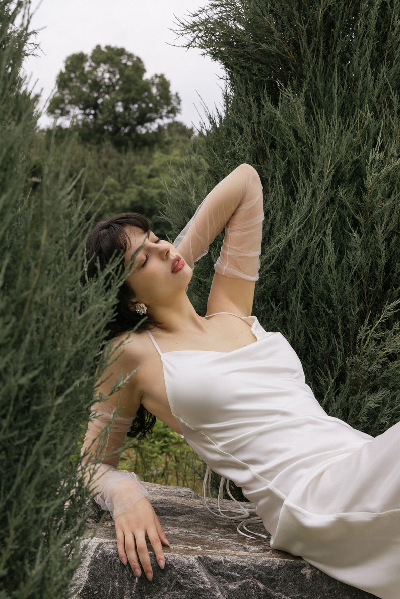 May include: A woman wearing a white satin dress with a cowl neck and thin straps. She is wearing sheer white gloves and has her eyes closed. She is sitting on a rock with greenery behind her.