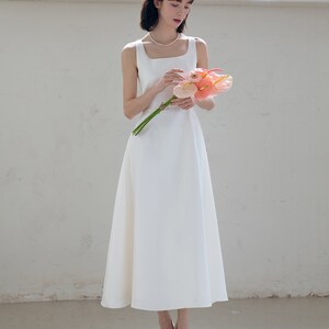 May include: A white sleeveless midi dress with a square neckline and a fitted bodice. The dress has a flowing skirt and is worn with a white headband and white heels.