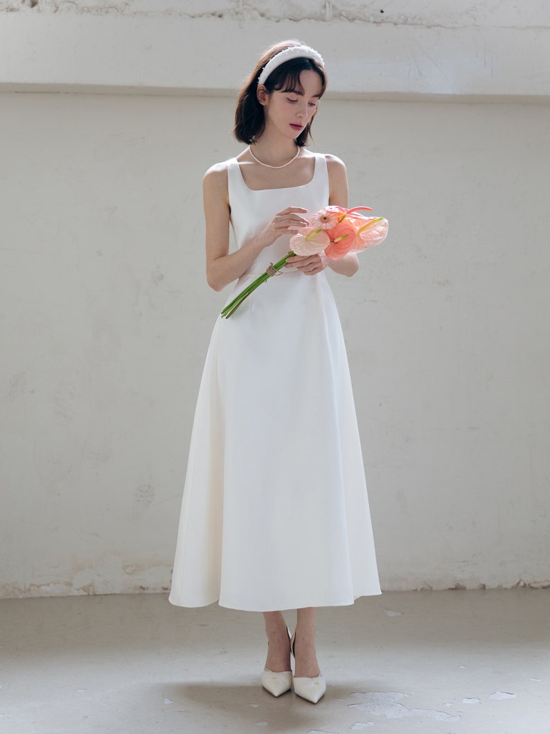 May include: A white sleeveless midi dress with a square neckline and a fitted bodice. The dress has a flowing skirt and is worn with a white headband and white heels.
