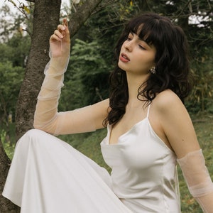 May include: A woman wearing a white satin dress with a cowl neck and sheer mesh gloves sits on a tree branch. She is holding a dandelion in her hand and looking up.