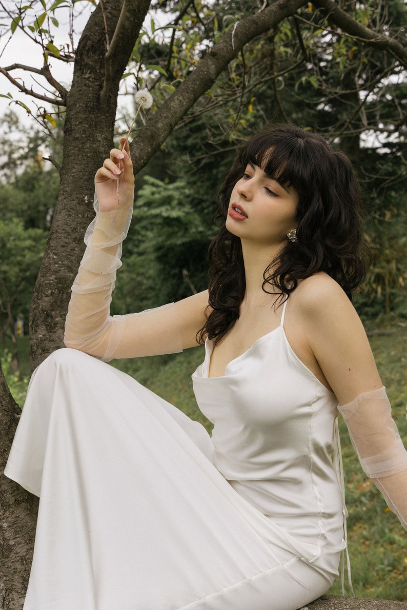 May include: A woman wearing a white satin dress with a cowl neck and sheer mesh gloves sits on a tree branch. She is holding a dandelion in her hand and looking up.