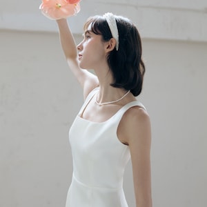 May include: A woman wearing a white sleeveless dress with a square neckline and a pearl necklace. She is holding a bouquet of pink flowers and wearing a white headband.