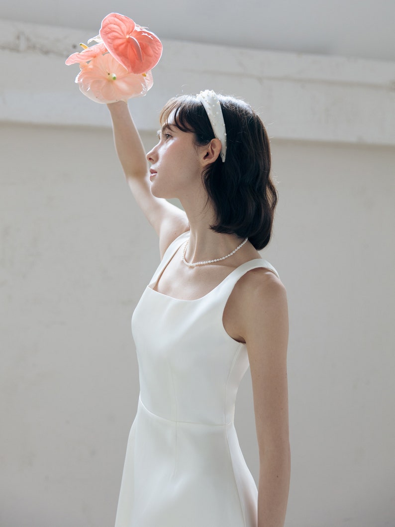 May include: A woman wearing a white sleeveless dress with a square neckline and a pearl necklace. She is holding a bouquet of pink flowers and wearing a white headband.