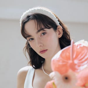 May include: A white headband with pearl embellishments, worn by a person with dark hair and a white sleeveless top. The person is holding a bouquet of pink flowers.