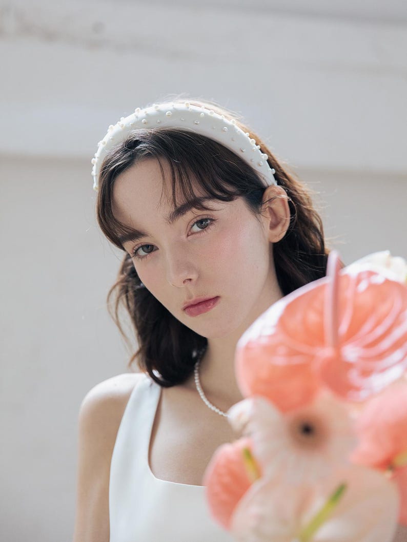 May include: A white headband with pearl embellishments, worn by a person with dark hair and a white sleeveless top. The person is holding a bouquet of pink flowers.
