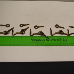 Op de afbeelding: Een verzameling messingkleurige haak- en oogsluitingen met bijpassende schroeven. De haken zijn gebogen en de ogen zijn cirkelvormig. Een groene liniaal met de tekst "Always be careful with fire" is aanwezig.
