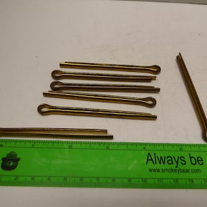 May include: Several gold-coloured split pins of different lengths, some with a loop at one end. A green ruler with the text "Always be careful" is in the foreground, measuring in inches.