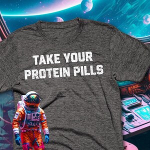 May include: A dark gray t-shirt with the text "TAKE YOUR PROTEIN PILLS" in white. An astronaut in an orange spacesuit is in the lower left corner of the image.
