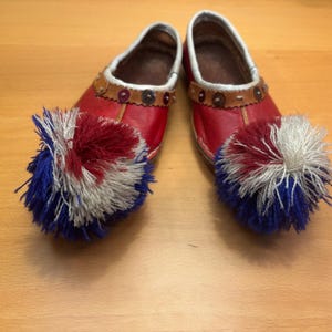 May include: A pair of red leather shoes with white trim and decorative brown leather straps. Each shoe features a large pom-pom in red, white, and blue. The shoes are displayed on a wooden surface.