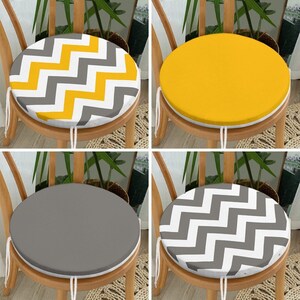 Può includere: Quattro cuscini rotondi per sedie con diversi motivi. Uno presenta un motivo a zig-zag giallo, bianco e grigio. Un altro è giallo tinta unita. Un terzo è grigio tinta unita e l'ultimo ha un motivo a zig-zag bianco e grigio. Ogni cuscino è legato alla sedia.
