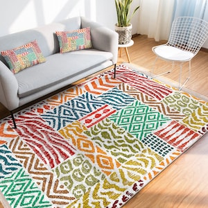 Op de afbeelding: Een kleurrijk vloerkleed met een patchwork-ontwerp met geometrische patronen in tinten rood, oranje, geel, groen, blauw en bruin. Het kleed ligt op een houten vloer in een woonkamer.