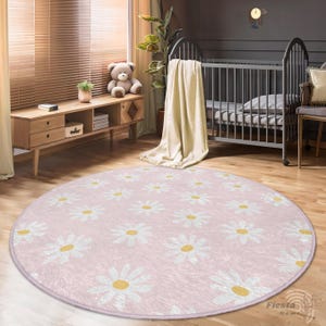May include: A pink round rug with a white daisy pattern. The rug is in a nursery with a crib and a wooden dresser.