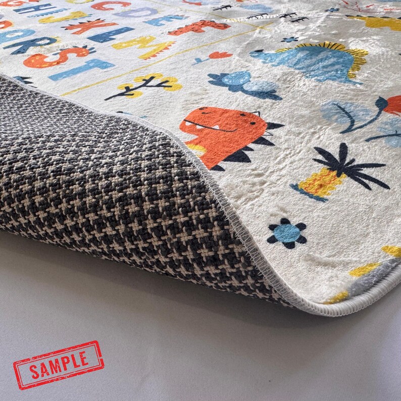 May include: A white children's rug with colorful illustrations of dinosaurs, flowers, and the alphabet. The rug's underside is a dark gray and white patterned fabric. The rug is rectangular and has a gray border.