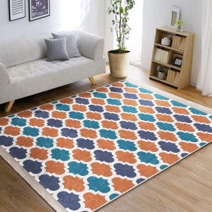May include: A rectangular area rug featuring a repeating quatrefoil pattern in shades of burnt orange, teal, and navy blue on a white background, finished with short white fringe along the two shorter edges, laid flat on a wooden floor.