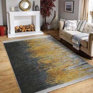 May include: A gray and gold area rug with a modern abstract design. The rug has a fringe border and is laid out on a hardwood floor in a living room setting.