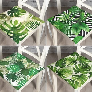 May include: Four square chair cushions with tropical leaf patterns. Each cushion features a different design with varying shades of green leaves on a white background. The cushions are tied to white chairs with white strings. The designs include monstera leaves and palm fronds.