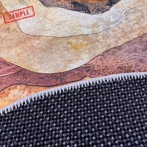 a close-up view of a woven rug or carpet with a black and gray pattern. The rug appears to be made of a textured material, possibly a synthetic fiber, and has a sample of the rug visible in the foreground.