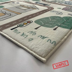 May include: A soft, off-white play mat featuring a whimsical city scene with roads, buildings, trees, and a traffic light. The mat has a stitched border and includes the text "No my da" and "A un um".