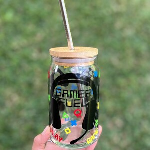 Puede incluir: Un tarro de vidrio transparente con un dise&ntilde;o negro de "Gamer Fuel", que incluye un auricular y s&iacute;mbolos de mandos de juego coloridos. El tarro tiene una tapa de bamb&uacute; y una pajita de acero inoxidable.