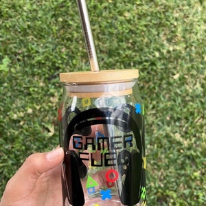 Puede incluir: Un tarro de vidrio transparente con tapa de bamb&uacute; y una pajita de acero inoxidable. El tarro tiene un dise&ntilde;o en blanco y negro con las palabras "Gamer Fuel" y s&iacute;mbolos de mando de juego de colores.