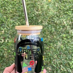 Puede incluir: Un tarro de vidrio transparente con tapa de bamb&uacute; y una pajita de acero inoxidable. El tarro tiene un gr&aacute;fico en blanco y negro de unos auriculares con el texto "Gamer Fuel" en &eacute;l. El tarro est&aacute; decorado con s&iacute;mbolos de mando de juego de colores.