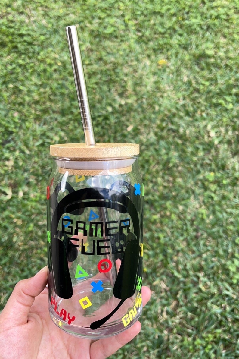 Puede incluir: Un tarro de vidrio transparente con tapa de bamb&uacute; y una pajita de acero inoxidable. El tarro tiene un gr&aacute;fico en blanco y negro de unos auriculares con el texto "Gamer Fuel" en &eacute;l. El tarro est&aacute; decorado con s&iacute;mbolos de mando de juego de colores.