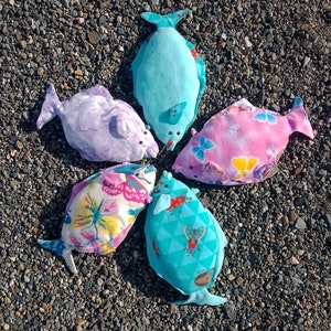 May include: Five colorful fabric fish toys arranged in a circle on a bed of small gray pebbles. The fish are made of different fabrics and have various patterns, including butterflies, geometric shapes, and solid colors.