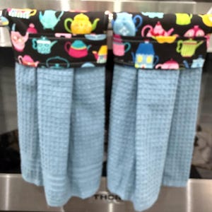 May include: Two light blue waffle-weave kitchen towels hanging side by side. Each towel features a black fabric top band decorated with a colorful pattern of various teapots in shades of yellow, pink, blue, and green, finished with a button closure.