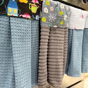 May include: A set of kitchen towels hanging on an oven. The towels are various shades of blue and gray, with patterned tops featuring teapots and floral designs. The oven is stainless steel.
