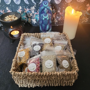 May include: A woven basket containing small, clear bags of dried herbs, each labelled with a white sticker. A black incense burner with a lit candle is next to a blue statue and a large lit candle. The background has a floral pattern.