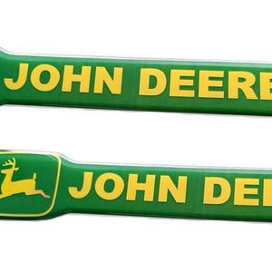 Könnte beinhalten: Zwei grün-gelbe John Deere Aufkleber mit einem stilisierten Hirsch-Logo.