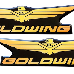 Könnte beinhalten: Zwei goldene und schwarze Motorrad-Embleme mit dem Text "GOLDWING" und einem stilisierten Adler mit den Buchstaben "GL" auf seiner Brust.