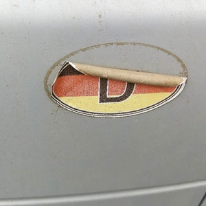 May include: A weathered oval sticker with the German flag colors of black, red, and yellow, and the letter "D". The sticker is peeling off a gray surface, likely a car.