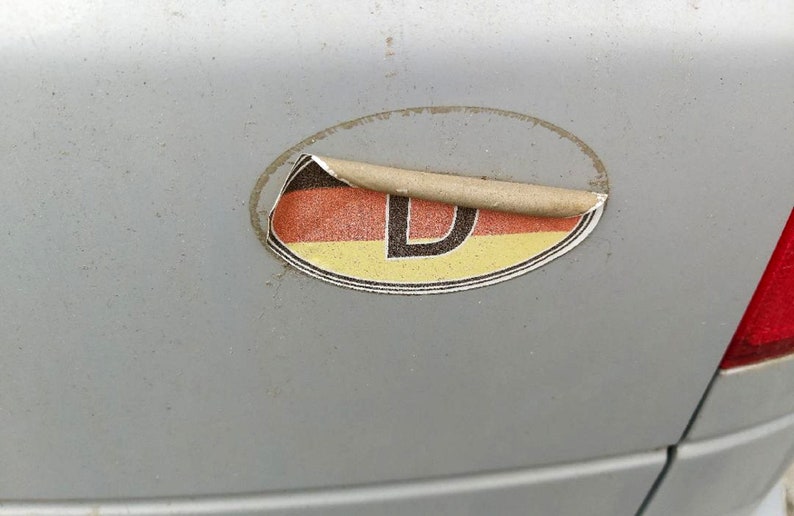 May include: A weathered oval sticker with the German flag colors of black, red, and yellow, and the letter "D". The sticker is peeling off a gray surface, likely a car.
