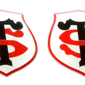Può includere: Due emblemi identici a forma di scudo. Ognuno presenta uno sfondo bianco con un design rosso e nero. Il design include una "T" nera e una "S" rossa intrecciate, con un bordo rosso.