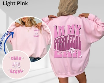 May include: Light pink sweatshirt with the text "In My Paralegal Era" in pink and white on the back. The front of the sweatshirt has a small design with the text "Para Legal" in pink and white.