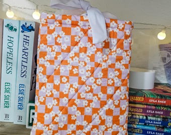 May include: A quilted book sleeve with a repeating pattern of orange and lavender checkerboard squares and white floral accents. The sleeve is tied with a white ribbon. The book sleeve is designed to protect books.